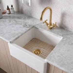 May include: A white farmhouse-style kitchen sink with a gold faucet and drain. The sink has a gold wire rack inside. The countertop is gray marble. The faucet has cross handles.