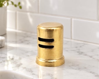 May include: A cylindrical gold-toned metal object with a smooth, polished finish. The item features two horizontal, parallel rectangular cutouts on its front side and a slightly flared base, presenting a simple, solid, and metallic construction.