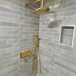 Puede incluir: Un juego de ducha dorado sobre un fondo de azulejos rectangulares grises. El juego incluye un cabezal de ducha de lluvia, una ducha de mano y un panel de control con manijas en forma de cruz. La ducha está integrada en una esquina con un estante empotrado.