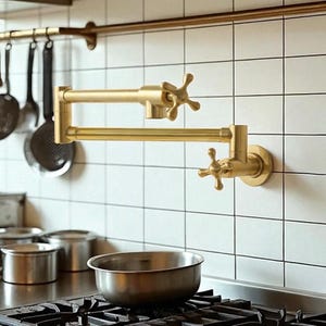 May include: A gold-colored, wall-mounted pot filler faucet with a cross-shaped handle. The faucet is installed on a white tiled wall above a stainless steel pot on a gas stove. Kitchen decor.
