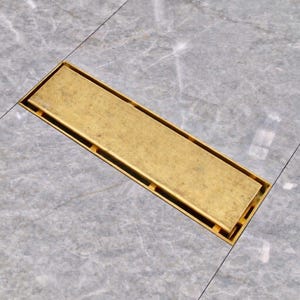 May include: A rectangular brass floor drain cover. The drain cover is a warm gold color and has a slotted design. The drain is set into a gray marble tile floor.