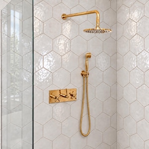 May include: A gold shower system featuring a wall-mounted rainfall showerhead, a handheld shower wand with a flexible hose, and a rectangular control plate with three cross-handle valves, all set against a wall of white hexagonal tiles.