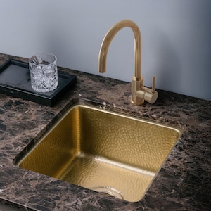 Op de afbeelding: Een goudkleurige gehamerde metalen spoelbak met bijpassende kraan. De spoelbak is verzonken in een donkerbruin marmeren aanrechtblad. Een decoratief glas en een zwart dienblad staan op het aanrecht.