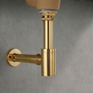 Puede incluir: Un accesorio de plomería dorado, probablemente una trampa en P, con un acabado pulido. El diseño incluye una tubería horizontal curva y una sección cilíndrica vertical. Montado en una pared de azulejos grises.
