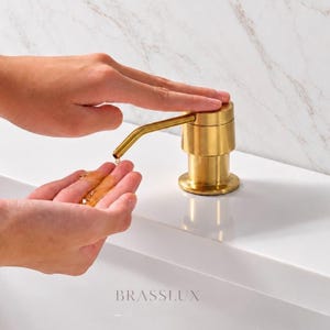 May include: A brushed gold soap dispenser with a cylindrical base and a horizontal spout. The dispenser features a metallic finish and is labelled with the text BRASSLUX at the base. It is designed for dispensing liquid soap into the palm of a hand.