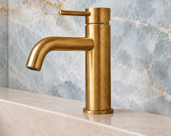 May include: A single-handle bathroom faucet with a brushed brass finish. The faucet features a cylindrical body and a curved spout, with a horizontal lever handle positioned at the top. The metal surface shows a textured, matte appearance.
