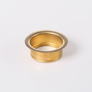 May include: A gold-colored sink drain with a circular design. The drain has a wide, flat rim and a cylindrical body with horizontal grooves. The interior is hollow, and the overall appearance is clean and modern.