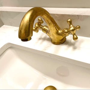 Peut inclure: Gros plan sur un robinet en laiton de style vintage avec des poignées en forme de croix. Le robinet a un bec courbé et est monté sur un comptoir blanc. Le luminaire est de couleur or chaud et semble vieilli.