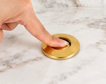 May include: A circular, brushed gold-toned metal pop-up drain stopper featuring a smooth, flat surface. The hardware has a metallic finish and a simple, round design that sits flush against the surrounding surface when in the closed position.