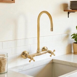 May include: A wall-mounted kitchen tap with a brushed brass finish, featuring a high-arched spout and two cross-style handles. The fixture is installed above a white sink against a white tiled backsplash, showcasing a classic design.