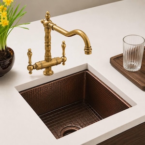 May include: A copper kitchen sink with a hammered texture, a brass tap, and a metal grid. A clear glass sits on a wooden tray. Yellow flowers in a brown pot are in the background.