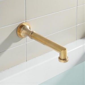 Könnte beinhalten: Ein goldfarbener Badezimmerhahn mit gebogenem Auslauf und kreisförmiger Basis ist an einer hell gefliesten Wand montiert. Der Wasserhahn befindet sich über einer weißen Badewanne.