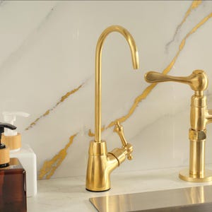 Puede incluir: Un grifo de cocina de oro cepillado con un ca&ntilde;o alto y curvado y una palanca lateral &uacute;nica. El grifo presenta un acabado met&aacute;lico y un dise&ntilde;o esbelto, colocado junto a una segunda manija de grifo dorada y un frasco dispensador de jab&oacute;n blanco.