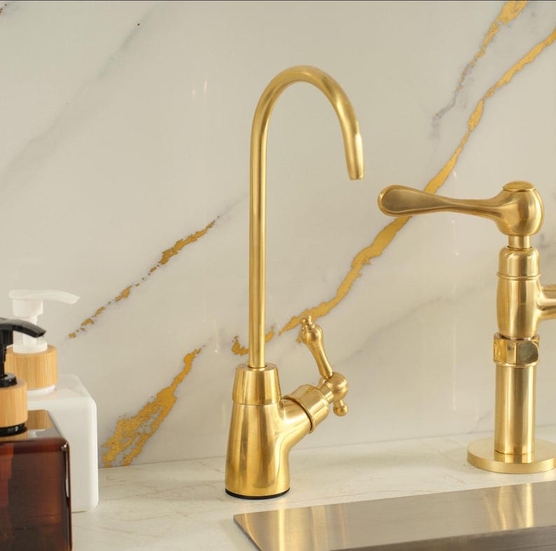 Puede incluir: Un grifo de cocina de oro cepillado con un ca&ntilde;o alto y curvado y una palanca lateral &uacute;nica. El grifo presenta un acabado met&aacute;lico y un dise&ntilde;o esbelto, colocado junto a una segunda manija de grifo dorada y un frasco dispensador de jab&oacute;n blanco.