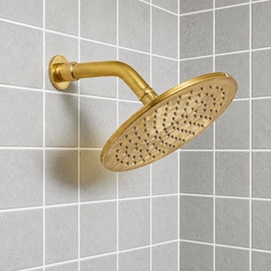 May include: A brushed gold shower head with a circular face and numerous small water nozzles. The shower head is mounted to a wall and features a curved arm connecting the circular head to the base, which is attached to a gray tiled wall.