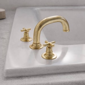 Puede incluir: Un grifo de baño dorado con un caño curvo y dos manijas en forma de cruz. El grifo está montado sobre una superficie blanca, probablemente un lavabo o encimera. El diseño es clásico y elegante.