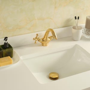 May include: A gold-coloured tap with cross handles is mounted on a white sink. A gold drain stopper is visible. A white sink basin is in the foreground. A green soap dispenser and a bar of soap are on the left. A white cup with toothbrushes is on the right.