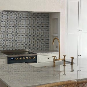 Puede incluir: Una isla de cocina con encimera de piedra gris claro y grifo de puente de lat&oacute;n. Detr&aacute;s de la isla hay una estufa negra empotrada en un hueco con un salpicadero de azulejos con patrones grises y blancos y armarios blancos a la derecha.