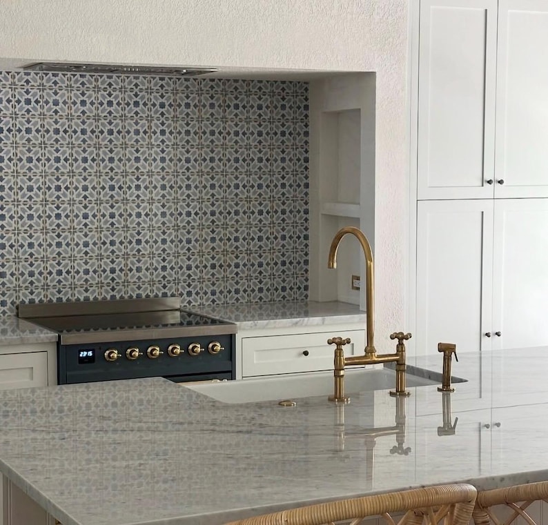 Puede incluir: Una isla de cocina con encimera de piedra gris claro y grifo de puente de lat&oacute;n. Detr&aacute;s de la isla hay una estufa negra empotrada en un hueco con un salpicadero de azulejos con patrones grises y blancos y armarios blancos a la derecha.