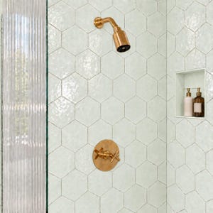 Puede incluir: Un área de ducha con azulejos hexagonales de color verde claro, un cabezal de ducha de latón y una válvula de control circular de latón a juego. Un nicho de pared empotrado contiene dos botellas de color ámbar con dispensadores dorados.