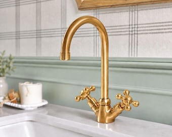 May include: A gold-toned metal faucet with a high-arched spout and two cross-shaped handles. The fixture features a smooth, polished finish and is mounted on a white countertop, showcasing a classic design with a curved neck and dual knobs.