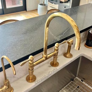 以下が含まれることがあります： 高いアーチ型の注ぎ口と2つのレバーハンドルを備えた、金色のキッチン蛇口。蛇口には、サイドスプレーと別の浄水器蛇口が含まれています。器具は白い大理石のカウンタートップに取り付けられています。