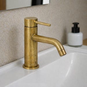May include: A gold-colored bathroom faucet with a curved spout and a lever handle. The faucet is mounted on a white sink, with a white and black soap dispenser in the background. The faucet has a cylindrical base and body.