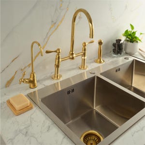 May include: A kitchen sink with a double basin and a gold-colored faucet set. The set includes a tall, arched faucet, a smaller faucet, and a soap dispenser. The sink is stainless steel, and the countertop is white with gray veining.