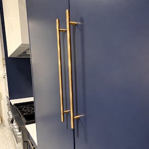 May include: A long, vertical, cylindrical brass cabinet handle with a polished metallic finish, mounted onto a matte navy blue cabinet door. The handle features two horizontal support posts that attach it securely to the flat, solid-colored surface.