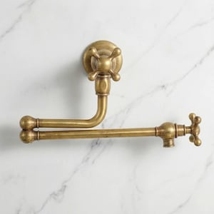 Puede incluir: Un grifo de llenado de ollas de estilo antiguo, montado en la pared, con acabado de latón. El grifo tiene un mango en forma de cruz y un caño curvo que se extiende horizontalmente. Fijado sobre una superficie con diseño de mármol blanco.