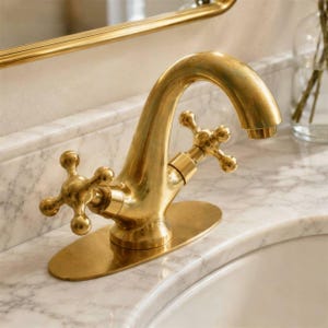 Puede incluir: Un grifo de baño de latón con un caño curvo y dos manijas en forma de cruz. El grifo está montado sobre una placa base ovalada y se coloca sobre una encimera de mármol blanco con un lavabo visible y un espejo con marco dorado arriba.