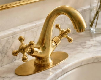 May include: A brass bathroom faucet featuring a curved spout and two cross-shaped handles. The faucet is mounted on an oval base plate and is positioned over a white marble countertop with a visible sink basin and a gold-framed mirror above.