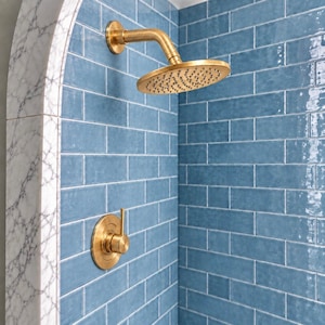 May include: A brushed gold shower head and a matching single-handle shower control valve mounted on a wall covered in glossy blue rectangular subway tiles, framed by a white marble arch with grey veining.