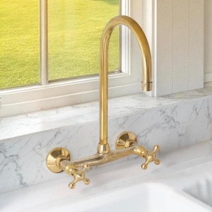May include: A wall-mounted kitchen tap with a brushed brass finish, featuring a high-arc spout and two cross-style handles. The fixture is installed against a white marble backsplash above a white sink, with a window visible in the background.