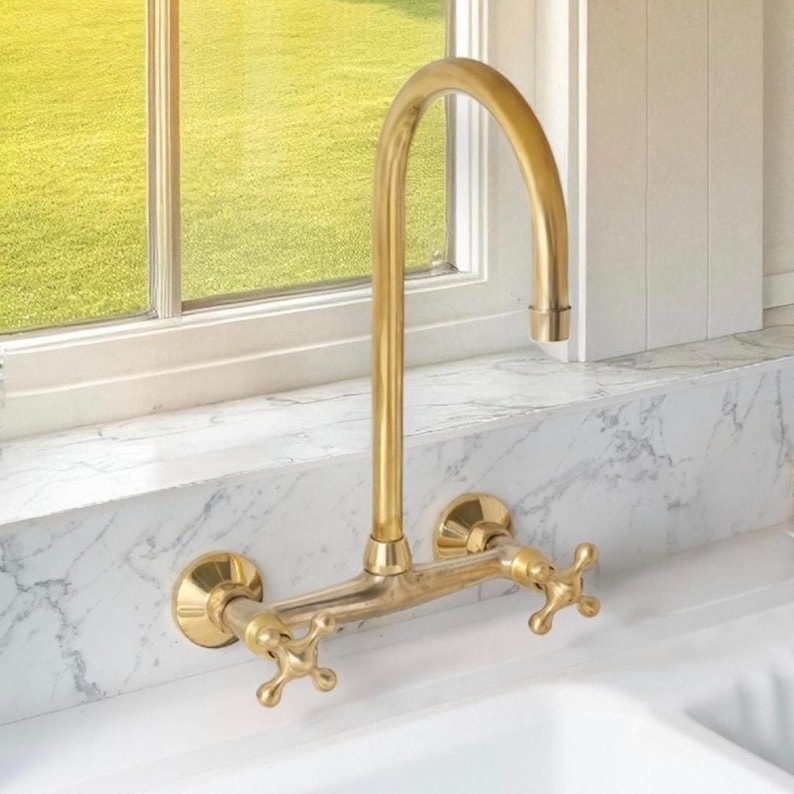 May include: A wall-mounted kitchen tap with a brushed brass finish, featuring a high-arc spout and two cross-style handles. The fixture is installed against a white marble backsplash above a white sink, with a window visible in the background.