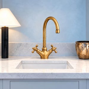 May include: A brass faucet with a curved spout and two cross-shaped handles, positioned above a white rectangular sink basin set into a white marble countertop. To the left is a black lamp with a white shade, and to the right is a textured gold bowl.