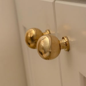 Puede incluir: Dos pomos de armario esféricos en tono dorado pulido con una superficie lisa y reflectante. Cada pomo presenta una cabeza redonda en forma de bola unida a una base cilíndrica, montada sobre una superficie de panel blanco con una costura visible.