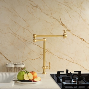 May include: A gold-coloured, articulated kitchen tap with a classic design. The tap is mounted on a white countertop, with a black hob and a plate of fruit in the foreground. The background features a marble-patterned wall.
