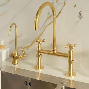 Puede incluir: Un juego de grifos de cocina en oro cepillado que incluye un grifo principal de arco alto con dos manijas en cruz y un grifo de agua filtrada m&aacute;s peque&ntilde;o. Ambos accesorios tienen un acabado met&aacute;lico y est&aacute;n montados sobre un fregadero de acero inoxidable.