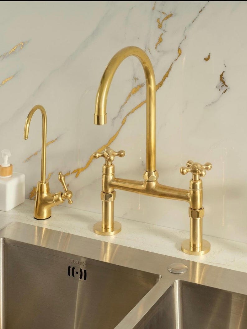 Puede incluir: Un juego de grifos de cocina en oro cepillado que incluye un grifo principal de arco alto con dos manijas en cruz y un grifo de agua filtrada m&aacute;s peque&ntilde;o. Ambos accesorios tienen un acabado met&aacute;lico y est&aacute;n montados sobre un fregadero de acero inoxidable.
