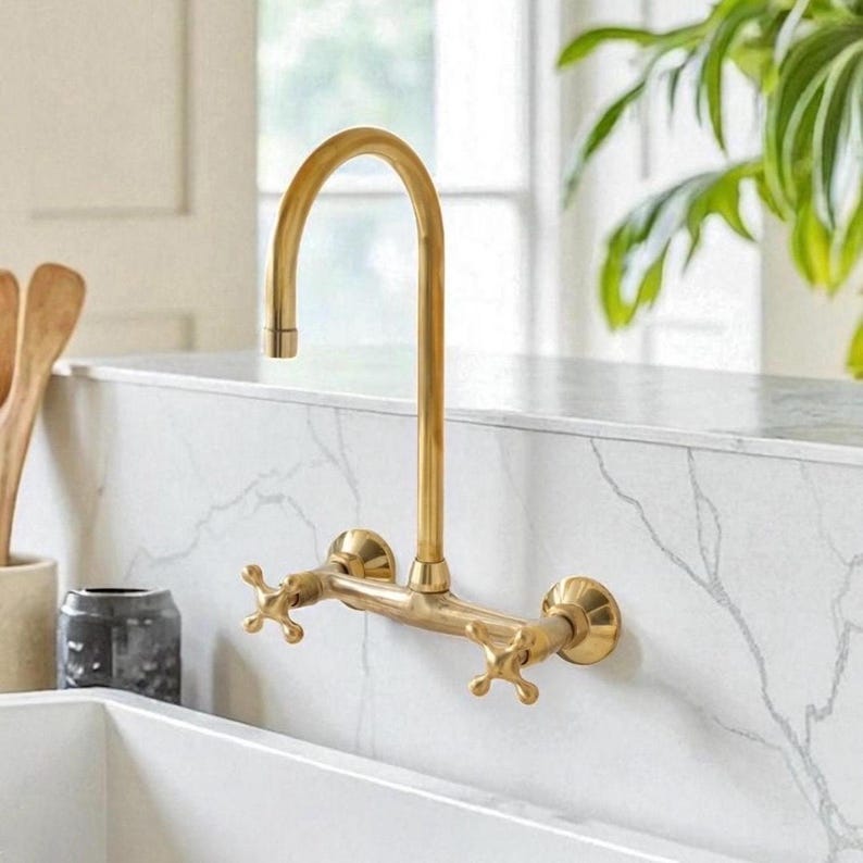 May include: A wall-mounted kitchen tap with a brushed gold finish, featuring a high-arc spout and two cross-style handles. The fixture is installed directly into a white marble backsplash with grey veining above a white farmhouse sink.