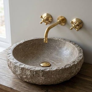 May include: A stone vessel sink with a rough, textured exterior sits on a wooden surface. The sink has a gold-colored drain cover. Above the sink, a gold faucet and two gold cross-handle fixtures are mounted on the wall.