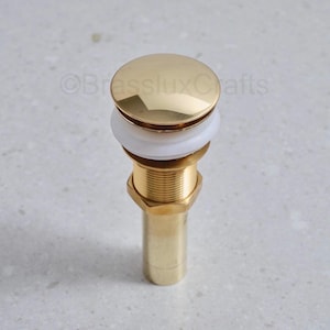 Puede incluir: Un tapón de desagüe de lavabo de color dorado con una parte superior redonda y plana y un cuerpo cilíndrico. El tapón tiene un anillo de goma blanco y una base roscada de color latón. El producto está diseñado para un lavabo de baño.