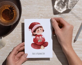May include: A greeting card featuring a watercolor illustration of a child in a red pajama set and hat. The card reads "HAVE A PAJAMA DAY" in black text. The card is held by hands.
