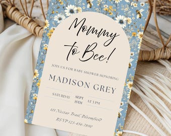 May include: A baby shower invitation with a floral and bee theme. The invitation features the text "Mommy to Bee!" in elegant script, with details for Madison Grey's baby shower. The background is a light blue with floral and bee illustrations.