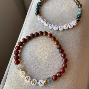 May include: Two beaded bracelets. One bracelet has the word "CREATURES" spelled out in white letter beads with gold lettering. The other bracelet has the word "DAWN" spelled out in white letter beads with gold lettering. The bracelets are made of various colored beads.