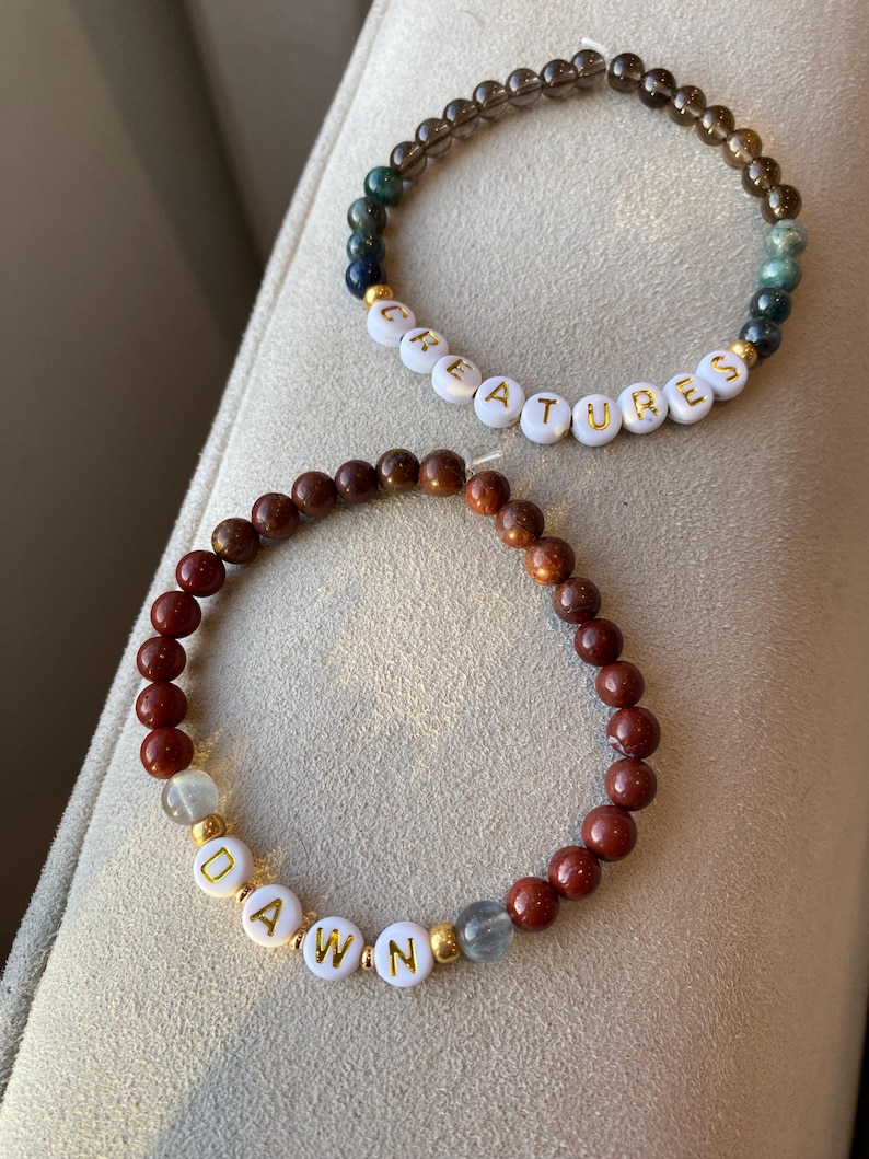 May include: Two beaded bracelets. One bracelet has the word "CREATURES" spelled out in white letter beads with gold lettering. The other bracelet has the word "DAWN" spelled out in white letter beads with gold lettering. The bracelets are made of various colored beads.
