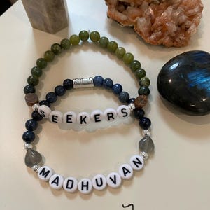 May include: Two beaded bracelets with the words "SEEKERS" and "MADHUVAN" spelled out in white letter beads. The bracelets are 7 inches in length. One bracelet has green beads, and the other has blue beads. Other crystals and stones are in the background.