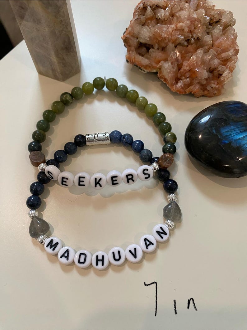 May include: Two beaded bracelets with the words "SEEKERS" and "MADHUVAN" spelled out in white letter beads. The bracelets are 7 inches in length. One bracelet has green beads, and the other has blue beads. Other crystals and stones are in the background.