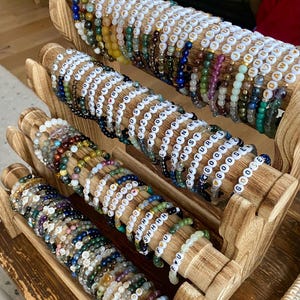 May include: A wooden display rack holds numerous beaded bracelets. The bracelets feature a variety of colorful beads and white letter beads spelling out words. The rack is made of light-colored wood, and the bracelets are arranged in neat rows.